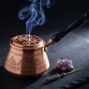 Puede incluir: Un quemador de incienso de cobre con un mango negro y patrones decorativos. Humo azul se eleva desde la parte superior. Un cristal de amatista y una ramita de lavanda están junto al quemador.