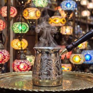 May include: A silver-toned metal coffee pot with an intricate embossed floral pattern and a long, dark wooden handle. The pot sits on a round, polished brass tray, with visible steam rising from the top opening of the vessel.