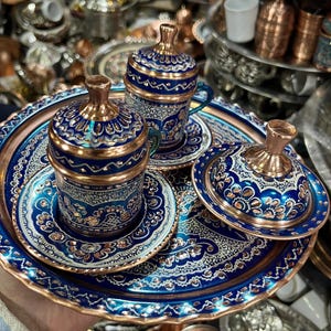 May include: A set of two copper coffee cups with matching saucers and a lidded sugar bowl, all arranged on a round serving tray. Each piece features a deep blue enamel finish with intricate silver-toned floral engravings and polished copper accents.