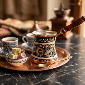 Può includere: Un set di caffè turco tradizionale su un vassoio di rame. Il set comprende una caffettiera decorativa, due tazzine, una ciotola di delizie turche e un cucchiaio. La caffettiera è nera con disegni floreali e un manico in legno.