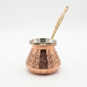 May include: A copper coffee pot with a wide base and a narrow neck, featuring intricate hand-engraved floral patterns on the exterior. The pot has a silver-toned interior lining and a long, slender, gold-toned metal handle with ornate detailing.
