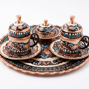 May include: A copper coffee set featuring two lidded cups with matching saucers and a small lidded bowl, all arranged on a large round tray. Each piece is decorated with black enamel and intricate hand-painted floral patterns in red, blue, and white.