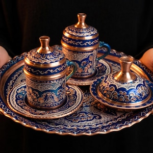 May include: A copper and blue Turkish coffee set, including two covered cups, saucers, and a serving tray. The set features intricate floral patterns and a metallic finish. The tray and cups are held by a person.