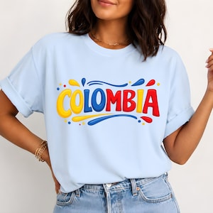 May include: A light blue short-sleeved t-shirt featuring the word COLOMBIA printed in a bold, rounded, multi-coloured font. The letters are yellow, blue, and red, accented with small colourful dots and fluid shapes surrounding the text.
