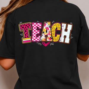 Pode incluir: Uma t-shirt preta com a palavra TEACH em letras grandes e coloridas nas costas. Cada letra tem um padrão único: estampado de leopardo rosa, xadrez rosa e branco, salpicos de tinta multicoloridos, smileys amarelos e papel pautado.