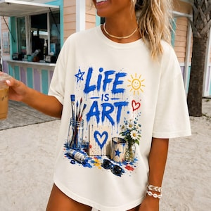 May include: An off-white t-shirt featuring a blue graphic print with the text "LIFE IS ART". The design includes a jar of paintbrushes, a canvas with a blue heart, a paint cup, a bouquet of daisies, a sun, a star, and black sunglasses.