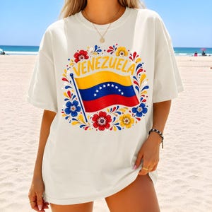 May include: An off-white short-sleeve t-shirt featuring a central graphic of the Venezuelan flag waving on a pole. The flag is surrounded by a circular arrangement of colorful red, yellow, and blue floral patterns with the word VENEZUELA in yellow.