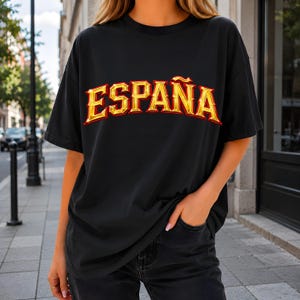 以下が含まれることがあります： 胸元に太字のアーチ状フォントで「ESPAÑA」という文字がプリントされた黒の半袖Tシャツ。文字はメタリックゴールド仕上げで赤い縁取りが施されており、暗い色の生地に対して立体的な効果を生み出しています。