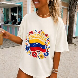 May include: An off-white short-sleeve t-shirt featuring a central graphic of the Venezuelan flag waving on a pole. The flag is surrounded by a circular floral pattern in red, yellow, and blue, with the word VENEZUELA arched above the flag.