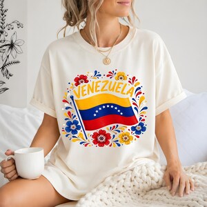May include: An off-white short-sleeve t-shirt featuring a graphic of the Venezuelan flag with the word VENEZUELA arched above it. The design is surrounded by a circular arrangement of stylized red, blue, and yellow floral patterns.