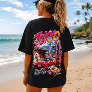 May include: A black t-shirt featuring a vibrant graphic print on the back. The design displays the word TOKYO in bold letters above a scene with a red sports car, a bowl of ramen, a lucky cat figurine, cherry blossoms, and city architecture.