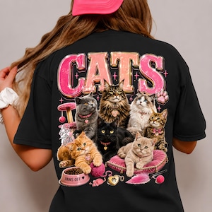 May include: A black t-shirt featuring a graphic print of seven cats in various colors. The text "CATS" appears in large pink letters at the top, with "Life is Better with Cats" and "PAWS OFF" printed on a pink food bowl at the bottom.