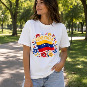 May include: A white short-sleeve t-shirt featuring a central graphic of the Venezuelan flag waving on a pole, surrounded by a circular arrangement of colorful red, yellow, and blue floral patterns with the word VENEZUELA printed in yellow.