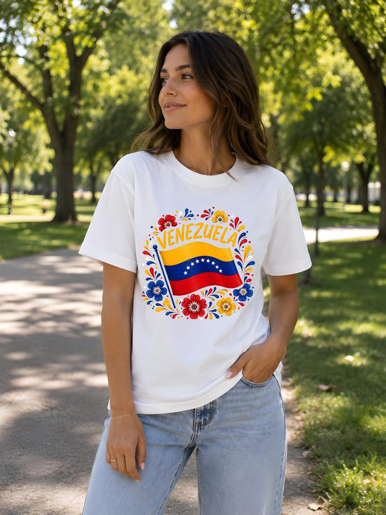 May include: A white short-sleeve t-shirt featuring a central graphic of the Venezuelan flag waving on a pole, surrounded by a circular arrangement of colorful red, yellow, and blue floral patterns with the word VENEZUELA printed in yellow.