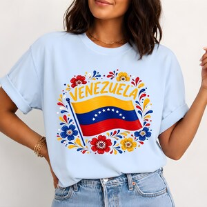 May include: A light blue short-sleeve t-shirt featuring a graphic print of the Venezuelan flag with the word VENEZUELA arched above it. The flag is surrounded by a circular decorative pattern of red, yellow, and blue floral motifs.