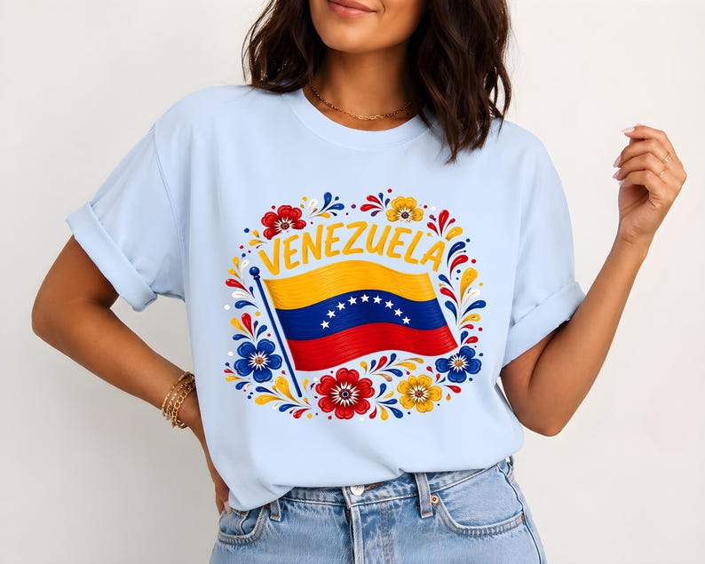 May include: A light blue short-sleeve t-shirt featuring a graphic print of the Venezuelan flag with the word VENEZUELA arched above it. The flag is surrounded by a circular decorative pattern of red, yellow, and blue floral motifs.