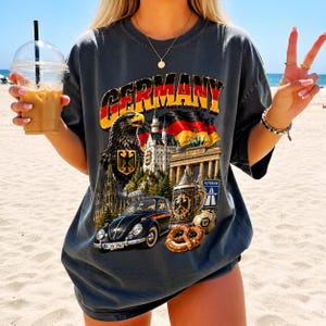 May include: A charcoal gray short-sleeve t-shirt featuring a graphic print with the word GERMANY in bold letters. The design includes a black eagle, a castle, the Brandenburg Gate, a vintage black car, a beer stein, a pretzel, and a soccer ball.