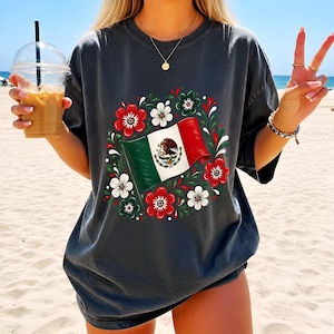 Puede incluir: Una camiseta gris carbón de manga corta con un gráfico central de la bandera de México ondeando, rodeada por una corona circular de flores rojas y blancas con hojas verdes. El diseño está estampado en la parte delantera de la prenda.