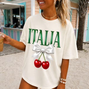 Può includere: Una t-shirt a maniche corte color bianco sporco con la parola ITALIA stampata in un audace carattere verde metallizzato. Sotto il testo c'è un'illustrazione grafica di due ciliegie rosso brillante appese a un grande fiocco bianco lucido.