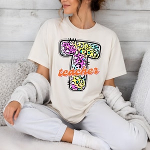 May include: A cream-colored short-sleeve t-shirt featuring a large letter T with a colorful leopard print pattern in shades of pink, blue, and yellow. The word teacher is written in a cursive orange font across the center of the letter T.