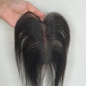 Peut inclure: Un complément capillaire composé de cheveux noirs lisses fixés sur une base en dentelle fine couleur chair. Les cheveux sont séparés par une raie centrale, laissant apparaître le maillage de la base qui imite l'aspect du cuir chevelu.