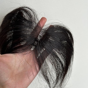 May include: A black hair extension piece featuring a fine mesh base with attached dark, straight hair strands. The hair is arranged in a layered, wispy style, designed to be clipped or attached to the scalp area for added volume or coverage.