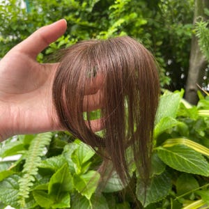 以下が含まれることがあります： ストレートのミディアムブラウンの毛束を使用したクリップ式ヘアエクステンション。前髪のスタイルで、滑らかな質感と自然な光沢があり、頭頂部に取り付けてボリュームを出したり、カバーしたりするように設計されています。