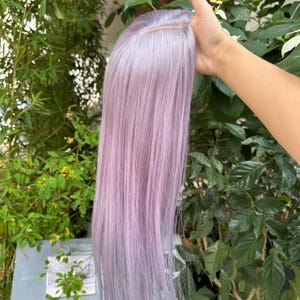May include: A long, straight wig featuring a soft pastel lavender color with a visible center part. The hair strands are smooth and sleek, extending downward in a uniform length, showcasing a light purple hue throughout the entire hairpiece.