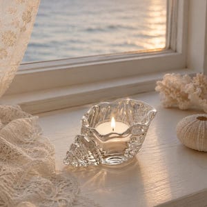Peut inclure: Un bougeoir en verre transparent en forme de coquillage, doté d'un design en spirale et d'un bord festonné. Une petite bougie chauffe-plat blanche est allumée à l'intérieur de la coquille en verre, diffusant une lueur chaude.
