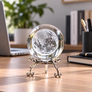 May include: A clear glass sphere containing a detailed 3D laser-etched moon model, resting on a decorative silver-toned metal tripod stand with ornate, curved legs. The sphere is transparent, showcasing the textured lunar surface inside.