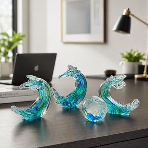 Puede incluir: Tres esculturas de vidrio con forma de olas rompiendo y un pisapapeles de vidrio esf&eacute;rico. Cada pieza presenta patrones de remolinos en vidrio azul, turquesa y transparente, con las esculturas de olas mostrando crestas curvas.