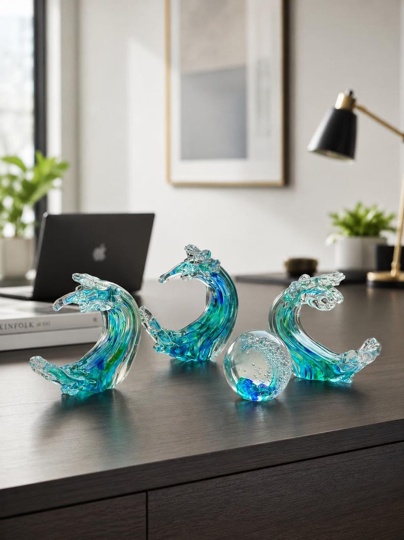 Puede incluir: Tres esculturas de vidrio con forma de olas rompiendo y un pisapapeles de vidrio esf&eacute;rico. Cada pieza presenta patrones de remolinos en vidrio azul, turquesa y transparente, con las esculturas de olas mostrando crestas curvas.