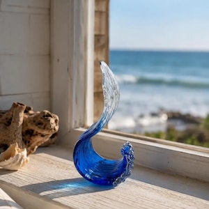 Puede incluir: Una escultura de vidrio con forma de ola oce&aacute;nica rompiendo, que presenta una base azul profundo que transiciona hacia una cresta transparente y texturizada. La pieza tiene una forma curva y fluida con detalles intrincados en los bordes.