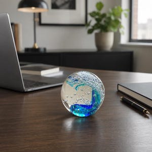 Puede incluir: Un pisapapeles de vidrio esf&eacute;rico con un exterior transparente y un dise&ntilde;o interno de cintas azules y verde azulado que se arremolinan, acentuado por numerosas burbujas de aire suspendidas en el material.