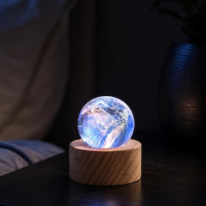 May include: A spherical glass orb featuring swirling blue, purple, and white nebula-like patterns illuminated from within, resting on a cylindrical light-colored wooden base with a visible grain texture and a flat top surface.