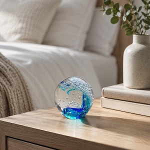 Puede incluir: Un pisapapeles esf&eacute;rico de cristal con un exterior transparente y un remolino interno de vidrio azul profundo y turquesa, acentuado por numerosas peque&ntilde;as burbujas de aire suspendidas en el centro de la esfera para crear un efecto de ola.