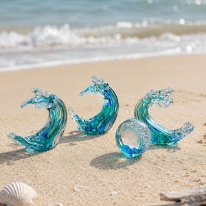 Puede incluir: Tres esculturas de vidrio con forma de olas del oc&eacute;ano rompiendo en tonos azules y verdes, acompa&ntilde;adas por una peque&ntilde;a esfera de vidrio transparente que contiene burbujas de aire y una base azul, dispuestas sobre arena cerca de la orilla.