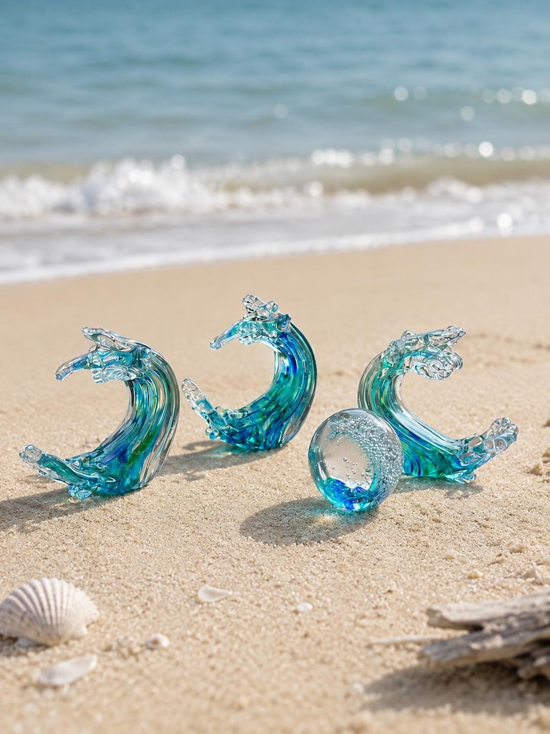 Puede incluir: Tres esculturas de vidrio con forma de olas del oc&eacute;ano rompiendo en tonos azules y verdes, acompa&ntilde;adas por una peque&ntilde;a esfera de vidrio transparente que contiene burbujas de aire y una base azul, dispuestas sobre arena cerca de la orilla.