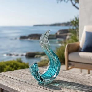 Puede incluir: Una escultura de vidrio transparente y azul verdoso con forma de ola estilizada. El objeto presenta una forma suave y curva con un degradado que va desde el azul verdoso intenso en la base hasta el vidrio transparente en la parte superior.