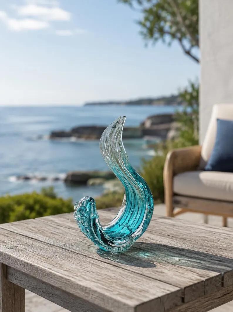 Puede incluir: Una escultura de vidrio transparente y azul verdoso con forma de ola estilizada. El objeto presenta una forma suave y curva con un degradado que va desde el azul verdoso intenso en la base hasta el vidrio transparente en la parte superior.