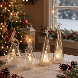 Puede incluir: Un conjunto de cuatro decoraciones de vidrio transparente que incluye dos adornos en forma de lágrima y dos adornos cónicos en forma de árbol. Cada pieza contiene un árbol blanco en miniatura iluminado, con una estrella en la punta de los conos.