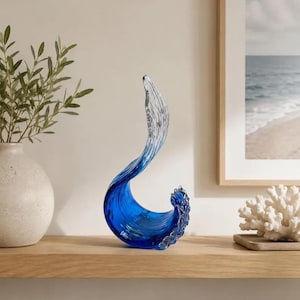Puede incluir: Una escultura decorativa de vidrio con forma de ola oce&aacute;nica rompiendo, que presenta una base azul intenso que se transforma en una parte superior clara y transl&uacute;cida. La pieza tiene una forma curva y fluida con detalles texturizados.