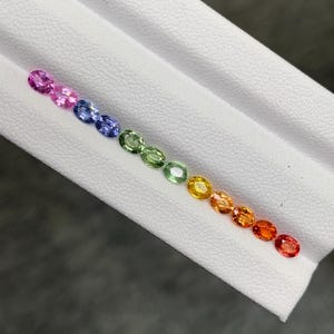 Puede incluir: Una colección de doce pequeñas piedras preciosas de corte ovalado dispuestas en una línea diagonal, mostrando un vibrante espectro de colores que incluye rosa, azul, verde, amarillo y naranja, sobre una superficie blanca texturizada.