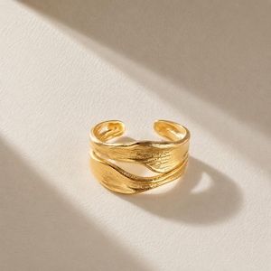 Op de afbeelding: Een goudkleurige verstelbare ring met een getextureerd, gelaagd ontwerp. De ring heeft een open ontwerp, waardoor de maat kan worden aangepast. Het oppervlak van de ring heeft een gerimpelde textuur, waardoor hij een unieke, organische uitstraling krijgt. De ring is waarschijnlijk gemaakt van metaal.