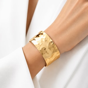 Peut inclure: Un large bracelet manchette doré doté d'une surface en métal martelé et poli, présentant une texture irrégulière et ondulée qui reflète la lumière sur toute la structure incurvée et solide du bracelet, offrant une finition très brillante.