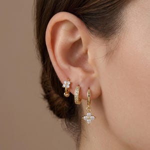 May include: Three gold hoop earrings worn in a single ear. The top earring features a small four-petaled flower charm with clear stones. The middle earring is a plain gold hoop with a row of clear stones. The bottom earring has a flower charm.