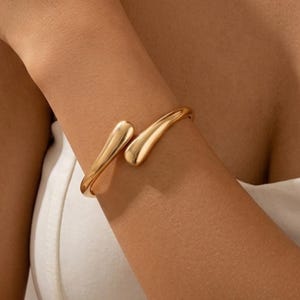 Op de afbeelding: Een goudkleurige metalen manchetarmband met een gladde, gepolijste afwerking. Het ontwerp bestaat uit een enkele doorlopende band die in een open stijl buigt, met twee afgeronde, druppelvormige uiteinden die elkaar in het midden tegenkomen.