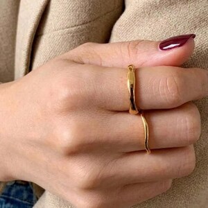 May include: Two gold-toned rings with a smooth, slightly irregular, organic surface texture. The rings are worn on the middle and ring fingers, featuring a polished finish that reflects light, creating a simple and clean appearance on the hand.