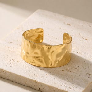 Può includere: Un ampio bracciale a fascia color oro caratterizzato da una superficie martellata, irregolare e altamente riflettente. Il bracciale presenta un design aperto con un bordo liscio e curvo, creando una forma continua e ondulata che cattura la luce.