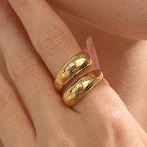 Puede incluir: Un anillo de tono dorado con un diseño de banda gruesa, pulida y redondeada. La superficie metálica es lisa y reflectante, creando un acabado de alto brillo que capta la luz. El anillo tiene un perfil curvo y sustancial alrededor del dedo.